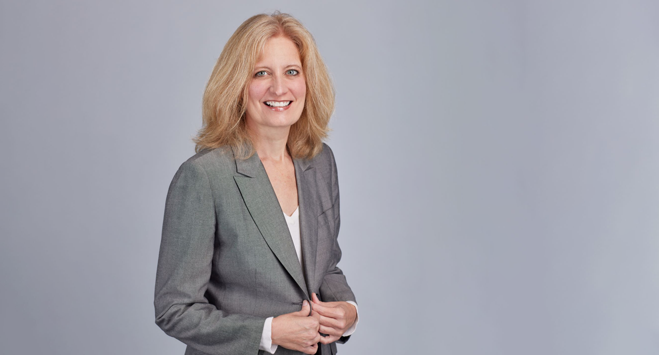 A blonde woman in a grey suit poses for a professional photo in front of a grey background.